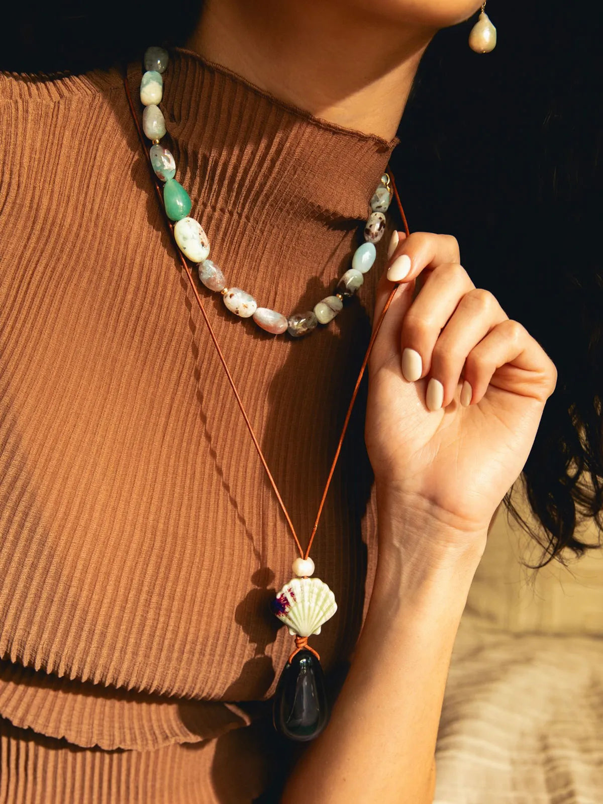 A close-up lifestyle shot of a woman wearing a bohemian-style layered necklace set. The set includes a short beaded necklace with green, white, and pink marbled beads, and a longer cord necklace featuring a white seashell pendant with a black teardrop stone and a small silver bead.