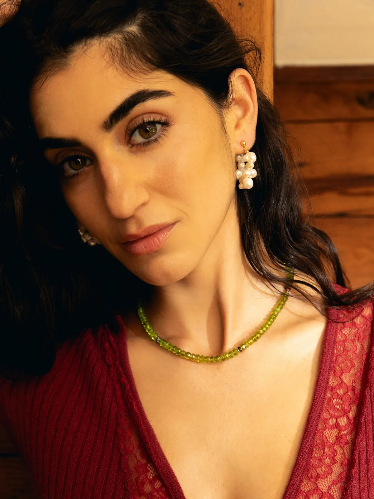A close-up portrait of a woman wearing a green beaded necklace and matching green and white flower-shaped earrings, paired with a deep red lace top.
