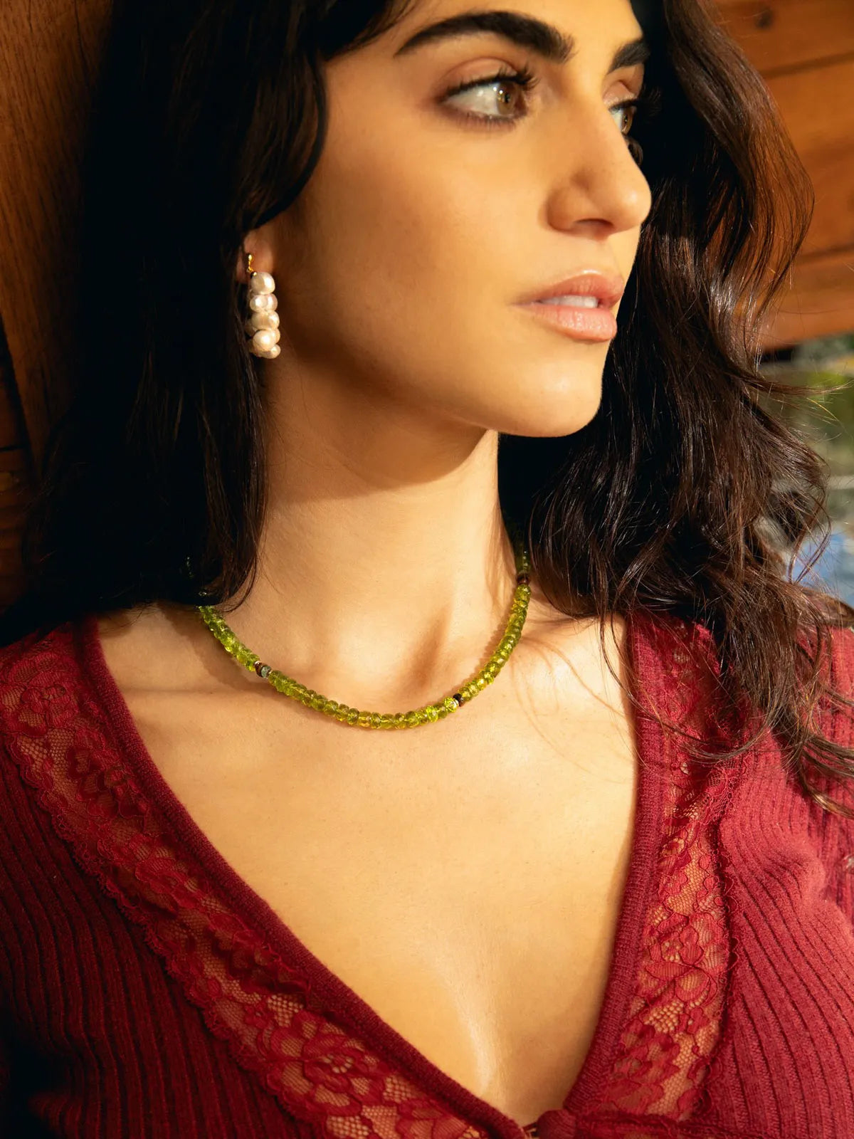 A close-up lifestyle shot of a woman wearing a green beaded necklace and matching drop earrings, paired with a deep red lace top.
