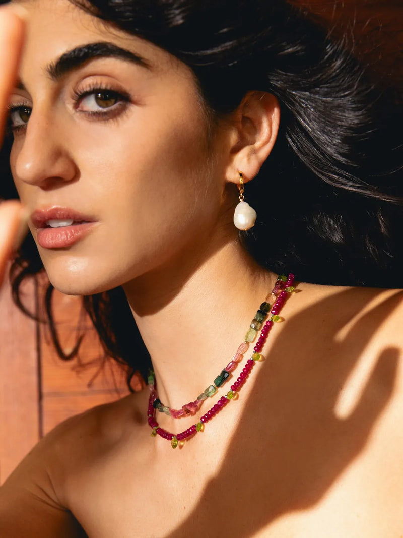 A close-up lifestyle shot of a woman wearing a multi-strand beaded necklace and matching drop earring. The jewelry features deep red, pink, green, and yellow beads with small gold star accents, set against a warm, sunlit background.