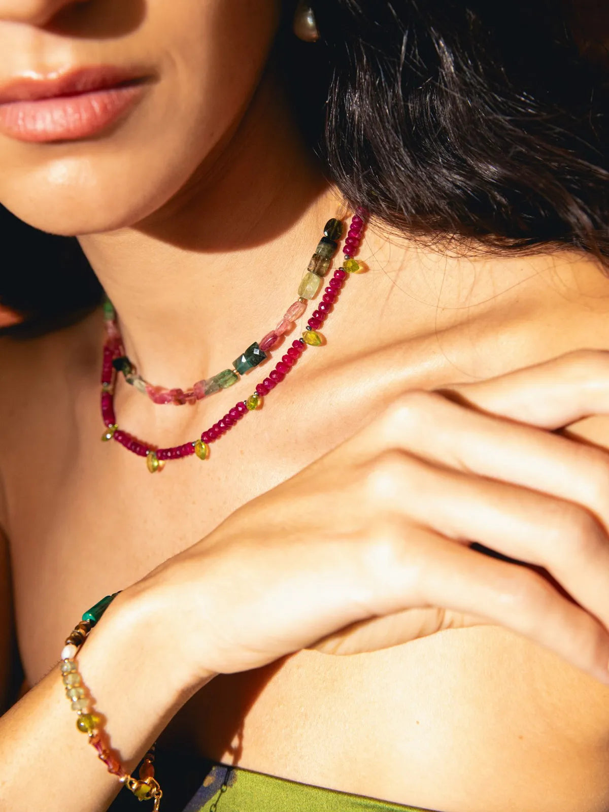 A close-up lifestyle shot of a model wearing a multi-strand beaded necklace and matching bracelet. The jewelry features colorful natural stone beads in pink, green, yellow, and brown, accented with small gold charms.