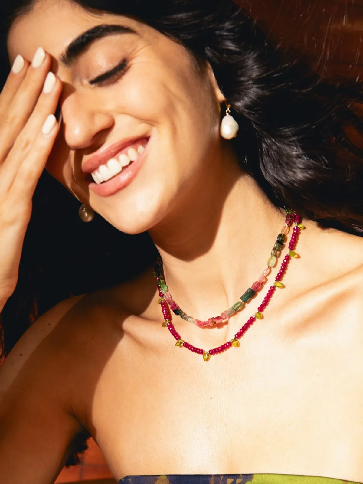 A close-up lifestyle shot of a smiling woman with dark hair, wearing a double-strand beaded necklace with red, green, and gold beads, and a matching pearl earring.