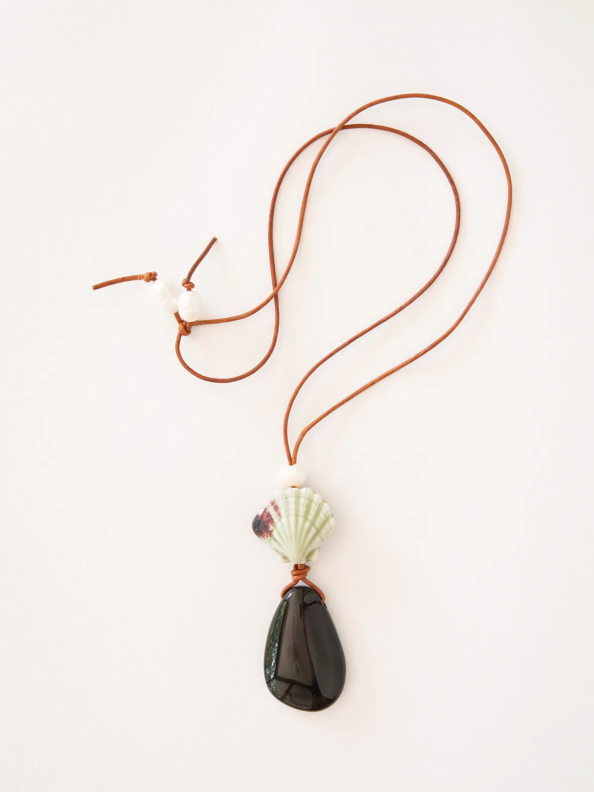 A necklace featuring a brown leather cord, a white seashell bead, freshwater pearls, and a dark teardrop-shaped stone pendant, displayed on a clean white background.