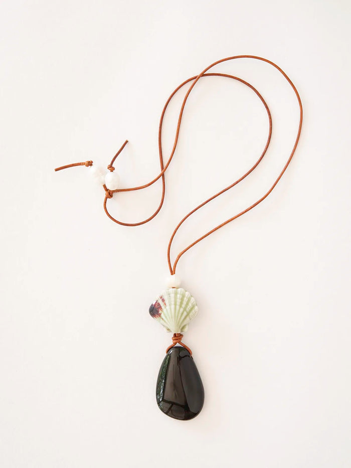 A necklace featuring a brown leather cord, a white seashell bead, freshwater pearls, and a dark teardrop-shaped stone pendant, displayed on a clean white background.
